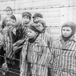 Child survivors of Auschwitz stand behind barbed wire fence wearing adult-sized prisoner jackets.