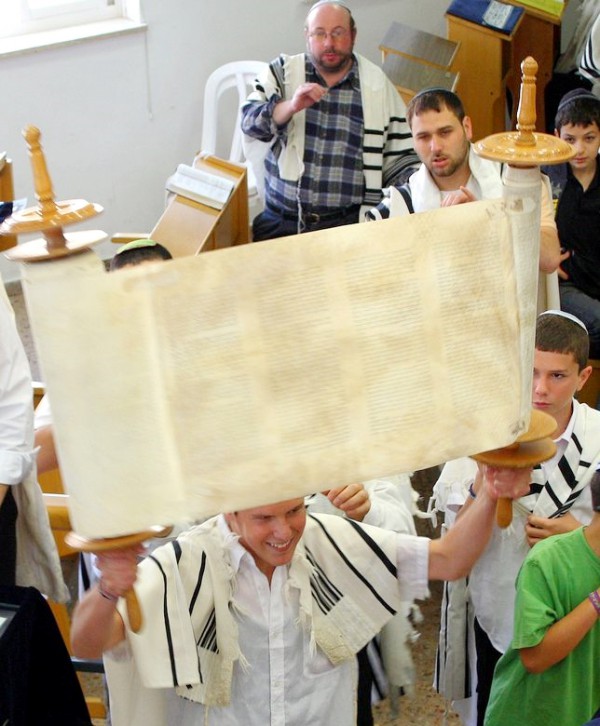 A Jewish man wearing tefillin (phylacteries) and a tallit (prayer shawl) holds  up the Torah scroll.