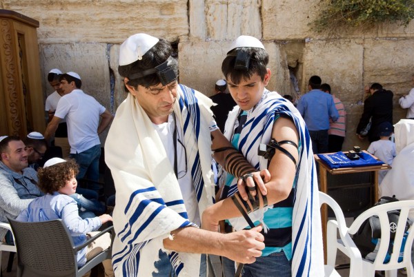 A Jewish man at the Western (Wailing) Wall helps a young man, perhaps his son, to wrap tefillin (phylacteries).