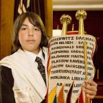 A Jewish teen holds a Torah scroll covered by an ornately decorated Torah mantle that commemorates those who perished in Nazi death camps during the Holocaust.