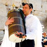 A Jewish man wearing tefillin (phylacteries) holds a Torah scroll protected by a study case called a tik.