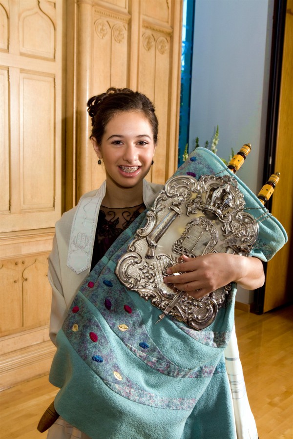 A Jewish girl holds the Torah scroll, which is covered by a Torah mantle and silver breastplate.