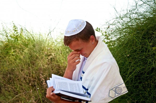 A Jewish teen wearing a tallit (prayer shawl) and a kippah (head covering) prays using a siddur (prayer book).