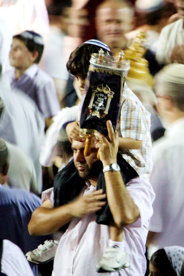 A father in the midst of a large crowd of people dancing and singing with the Torah carries his son on his shoulders as he parades a small Torah scroll on Simchat Torah. (Photo by Avital Pinnick)